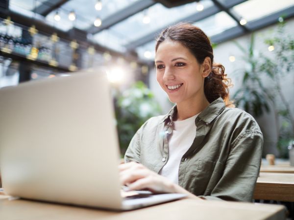 Una ragazza all'interno di un luogo pubblico che potrebbe essere un bar, arredato con luci appese e piante, sta usando il suo computer portatile al tavolo a cui è seduta e sorride mentre osserva lo schermo. Indossa una tshirt bianca con sopra una camicia verde militare aperta, ha i capelli castani lunghi raccolti in una coda.