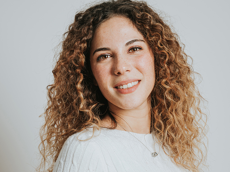 Michela Corazza, Account Manager VivaVox, fotografa di fianco su uno sfondo neutro: ha i capelli lunghi e ricci di colore castano chiaro e indossa una maglia di lana intrecciata leggera. Nella foto Michela sorride guardando in camera.