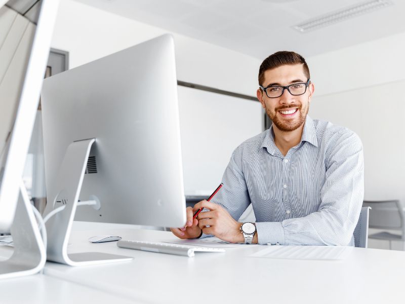 Un uomo seduto a una scrivania di fronte a un computer desktop: il suo ufficio è tutto bianco, lui indossa gli occhiali e una camcia chiara e sorride guardando in camera