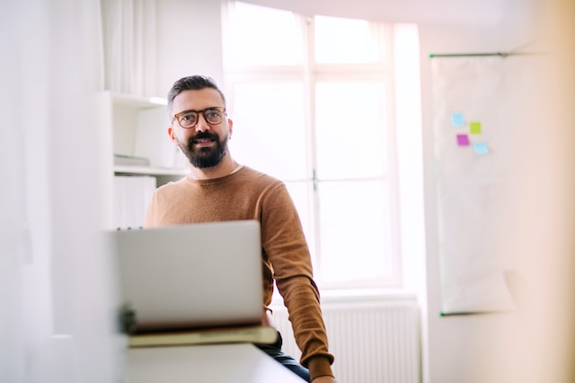 in una stanza illuminata dalla luce naturale che proviene da una finestra, un uomo siede davanti al suo computer portatibile e accenna un sorriso guardando l'obiettivo. L'uomo indossa gli occhiali da vista, un maglione marrone e ha capelli, barba e baffi scuri.